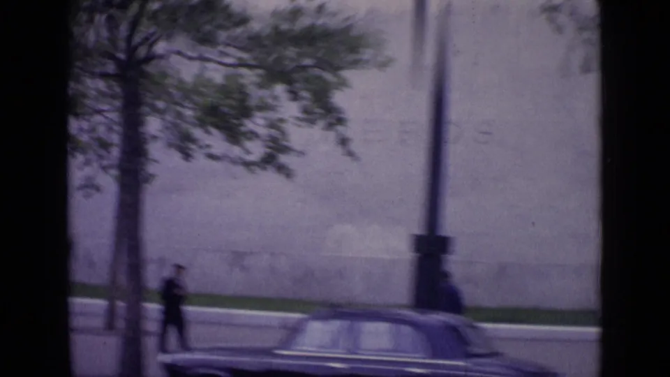 1962 Paris home-movie frame from a moving car showing trees, boulevard traffic, pedestrians, and a partial Eiffel Tower view.