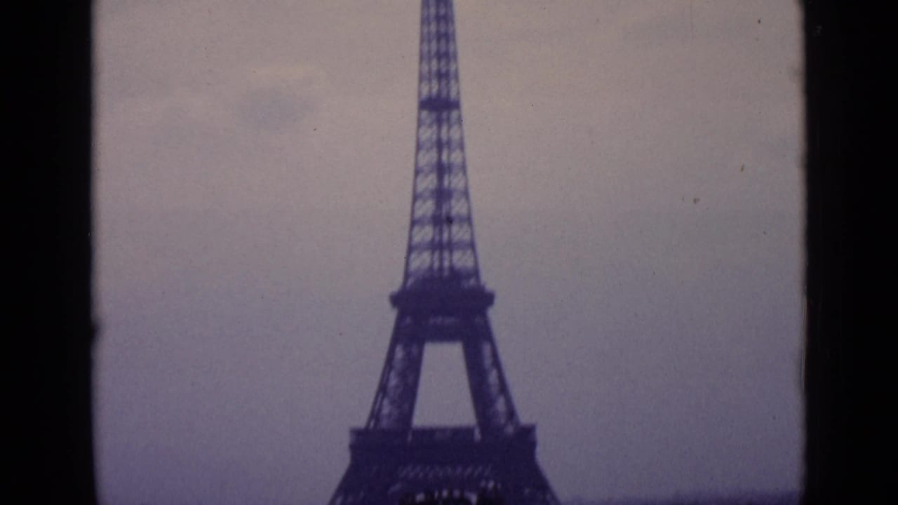 1962 Paris home-movie frame with the lower Eiffel Tower filling the view and a dark foreground divider crossing the shot.