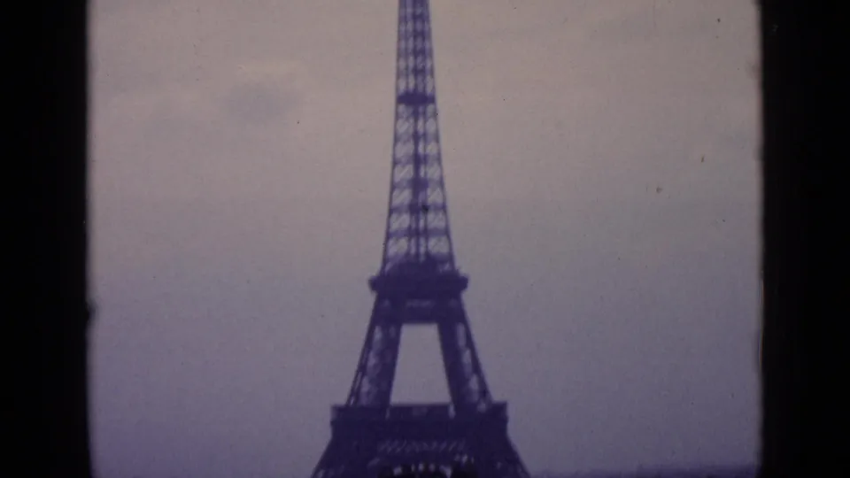 1962 Paris home-movie frame with the lower Eiffel Tower filling the view and a dark foreground divider crossing the shot.