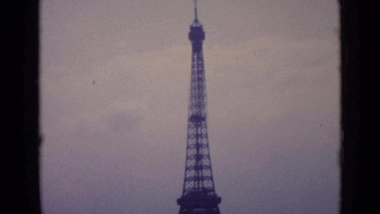 1962 Paris frame showing the Eiffel Tower centered at a distance, with soft grain, hazy sky, and dark home-movie frame edges.