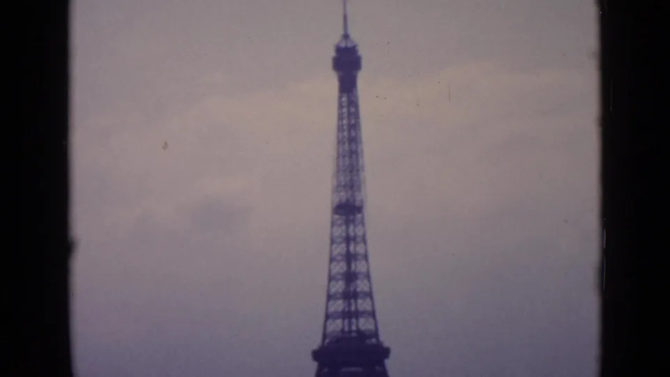 1962 Paris frame showing the Eiffel Tower centered at a distance, with soft grain, hazy sky, and dark home-movie frame edges.