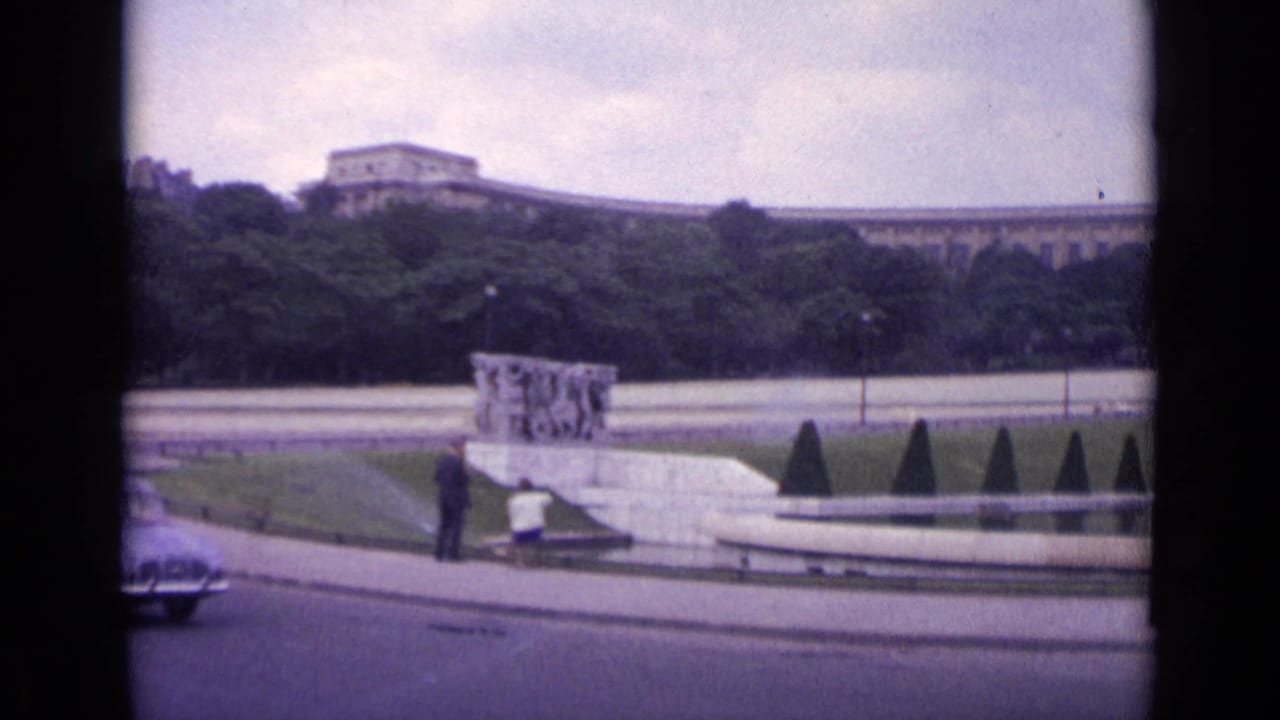Curved Paris roadway with a car passing people near a low sculptural monument and park landscaping.