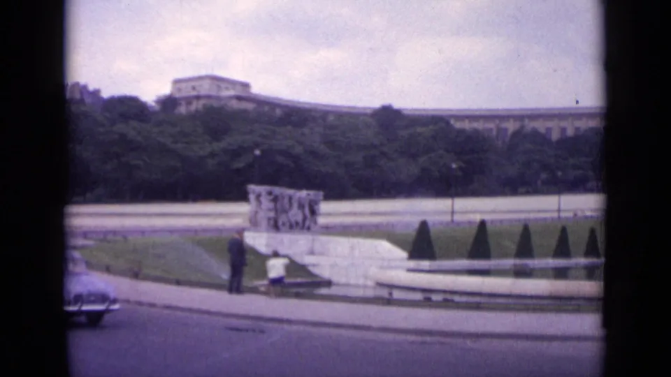 1962 Paris 8mm frame showing a curved road, a passing car, two distant people, benches, trimmed greenery, and a low sculptural monument.