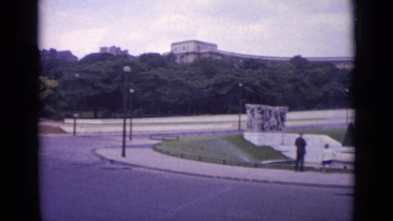 1962 Paris 8mm frame of a park-side roadway with one distant person near a low monument, benches, lampposts, trees, and buildings beyond.