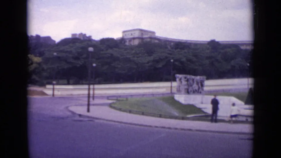 1962 Paris 8mm frame of a park-side roadway with one distant person near a low monument, benches, lampposts, trees, and buildings beyond.