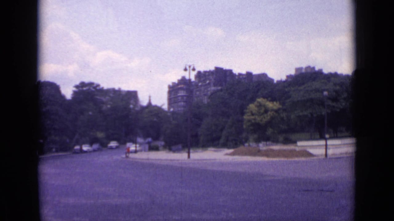 1962 Paris 8mm frame showing a broad street along a park, with small cars, lampposts, trees, and distant apartment buildings.