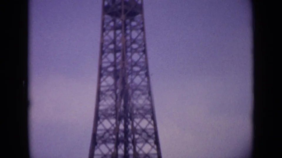 1962 8mm frame of the Eiffel Tower's narrowing lattice section in Paris, tinted purple with grain and soft exposure.