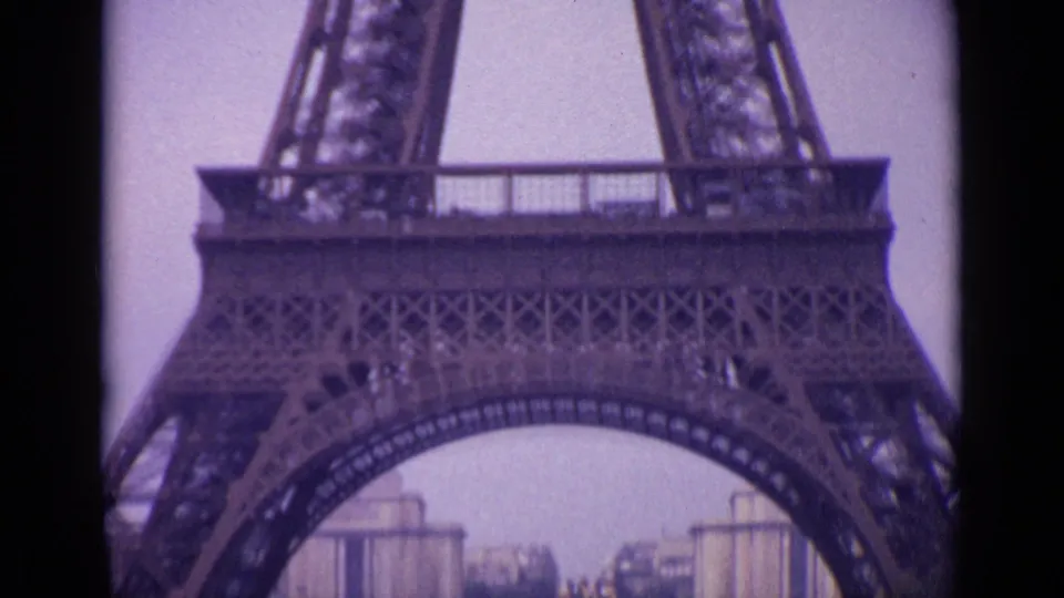 1962 Paris frame showing the Eiffel Tower base arch with distant buildings and tower ironwork seen through soft 8mm color.