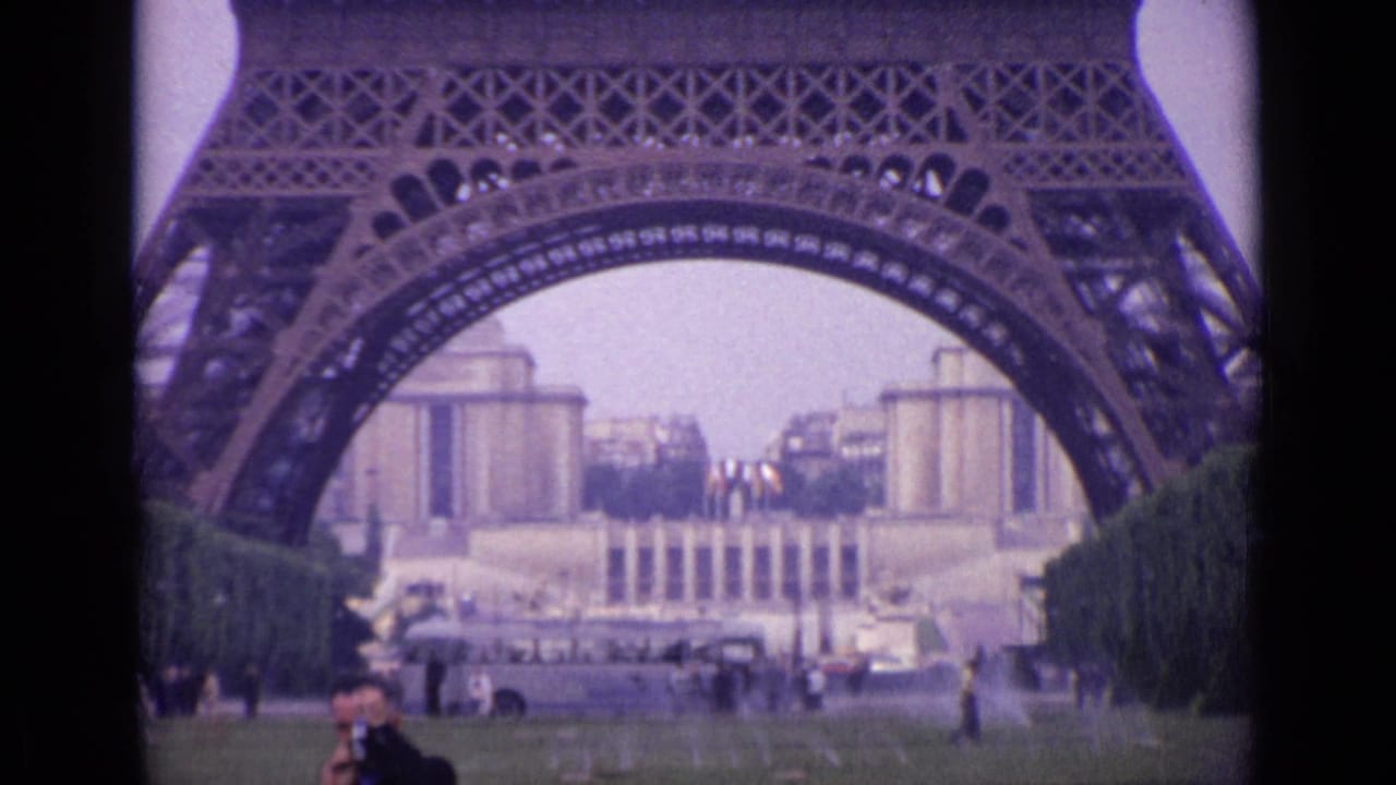 1962 Paris home-movie view of the Eiffel Tower base above Champ de Mars lawn, trees, pedestrians, and sprinklers.
