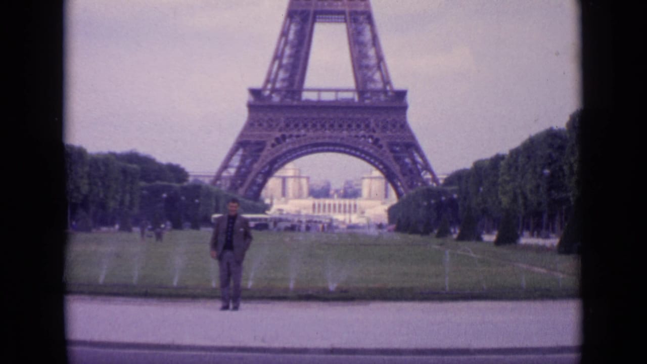 1962 Paris frame showing a man in a dark suit posed near lawn sprinklers with the Eiffel Tower in the background.
