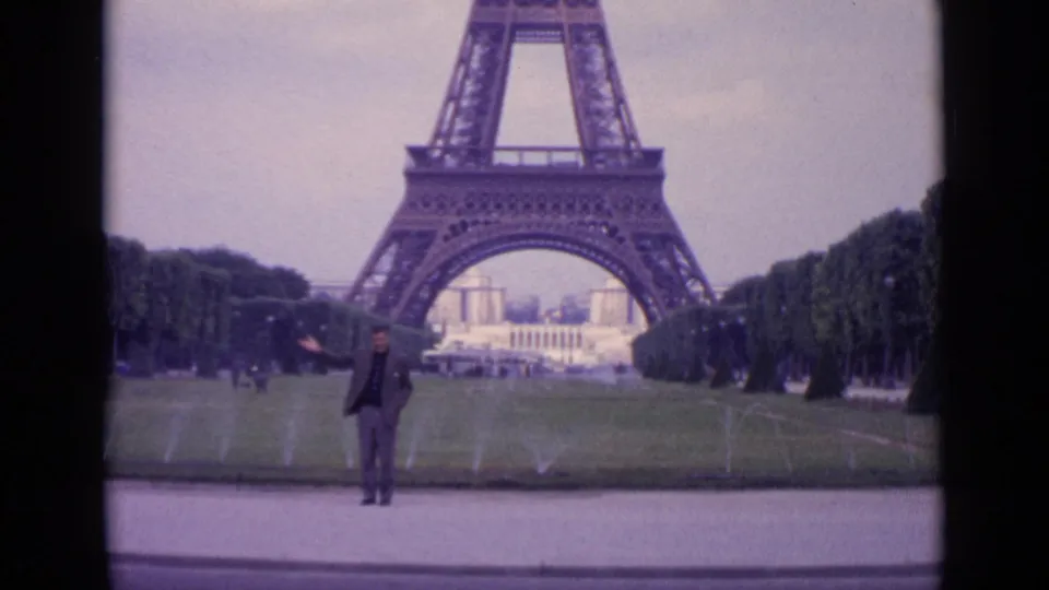 1962 Paris 8mm frame showing a man posing before the Eiffel Tower, with grass, fountains, trees, and cloudy sky visible.