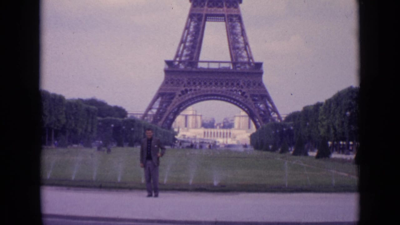 1962 Paris 8mm frame of a man posing beneath the Eiffel Tower, framed by lawns, fountains, trees, and overcast sky.