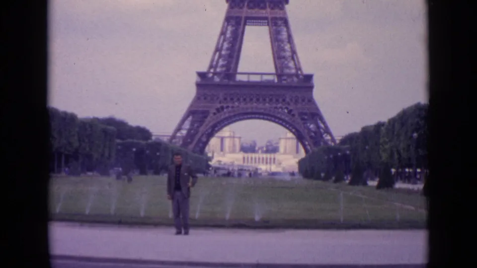 1962 Paris 8mm frame of a man posing beneath the Eiffel Tower, framed by lawns, fountains, trees, and overcast sky.