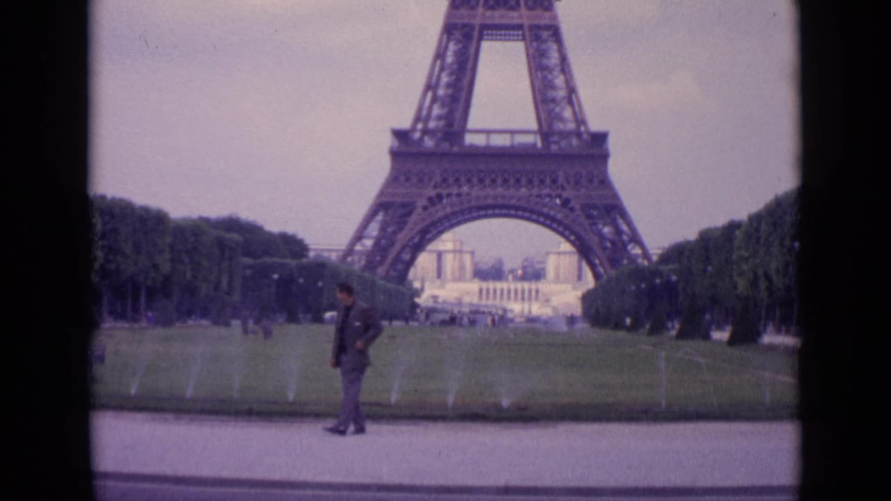 1962 Paris home-movie frame showing a suited man near a lawn and sprinklers with the Eiffel Tower base in the background.