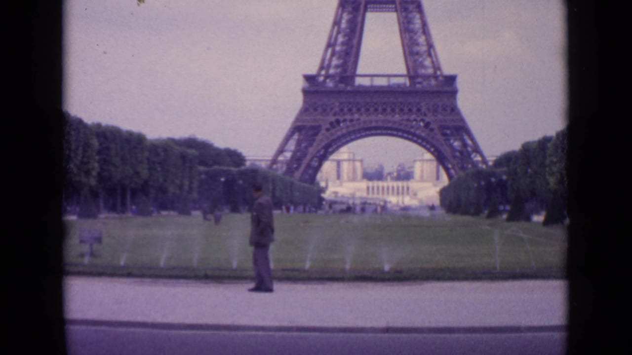 Suited man stands in profile near the lawn as sprinklers run below the Eiffel Tower.