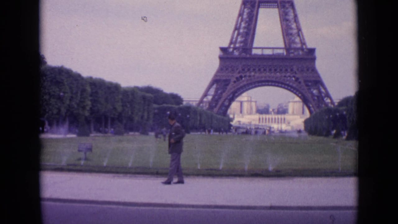 1962 Paris home-movie still showing the suited man closer to a lawn sign, with sprinklers and the Eiffel Tower base behind him.