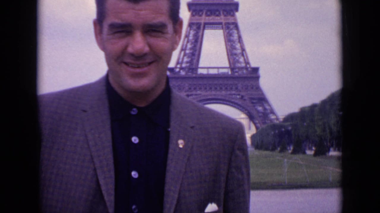 Close 1962 Paris home-movie portrait of a smiling man in a blazer, with the Eiffel Tower structure partly visible behind him.