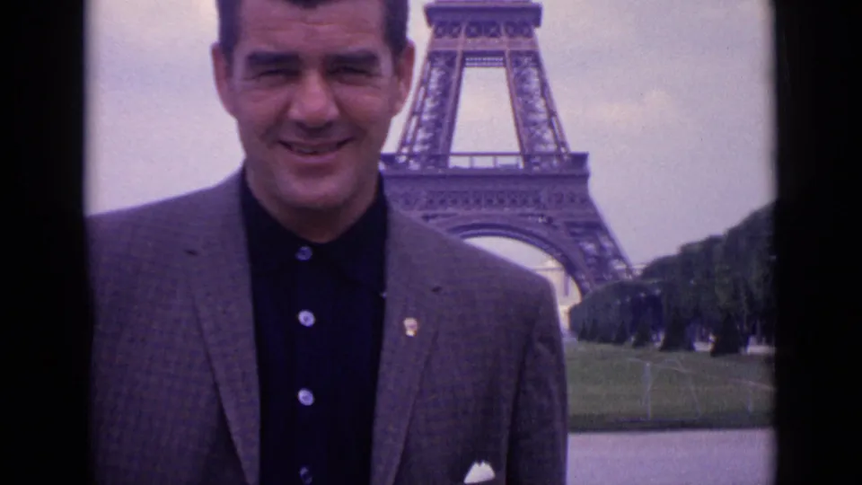 Close 1962 Paris home-movie portrait of a smiling man in a blazer, with the Eiffel Tower structure partly visible behind him.
