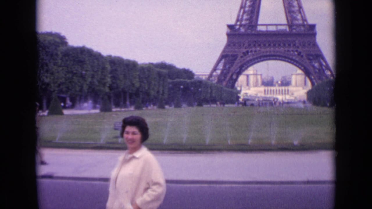 1962 Paris frame showing a woman in a light coat posing near the foreground with the Eiffel Tower, lawn, trees, and fountains behind her.