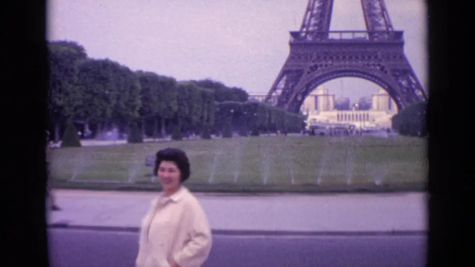 1962 Paris frame showing a woman in a light coat posing near the foreground with the Eiffel Tower, lawn, trees, and fountains behind her.