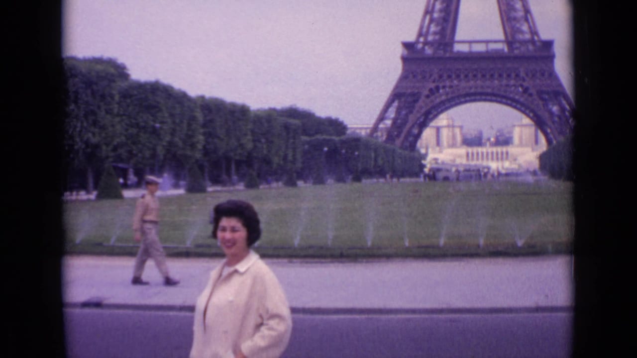 Woman in a light jacket stands smiling near the Eiffel Tower with lawns and fountains behind her.