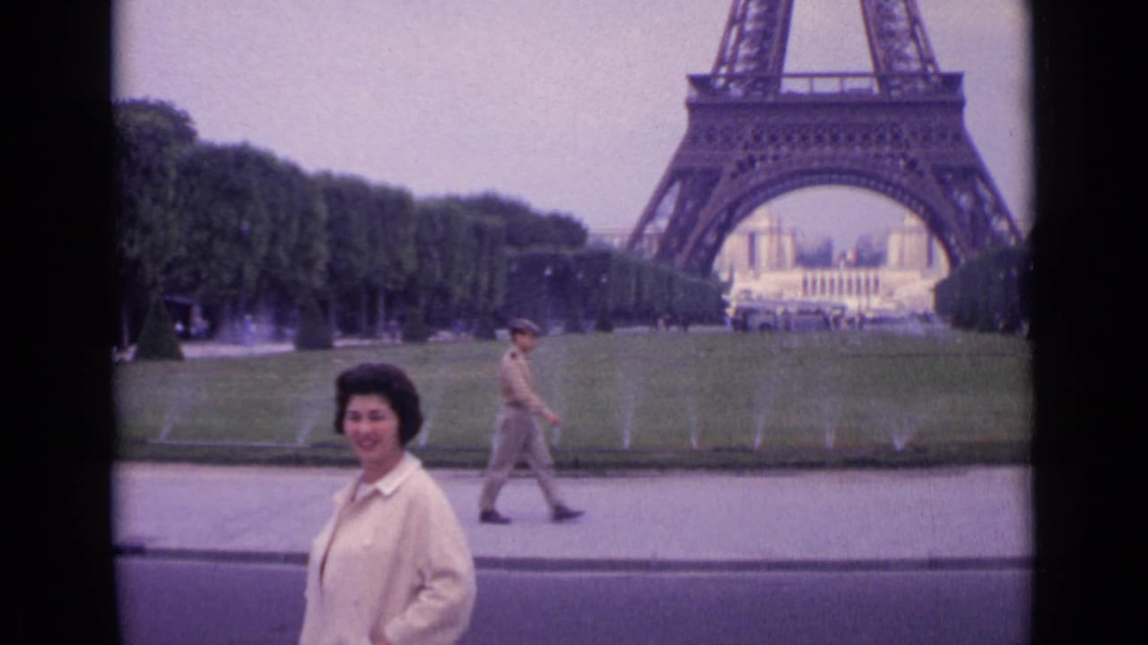 1962 Paris frame with a woman smiling in front of the Eiffel Tower while a uniformed passerby walks across the foreground path.