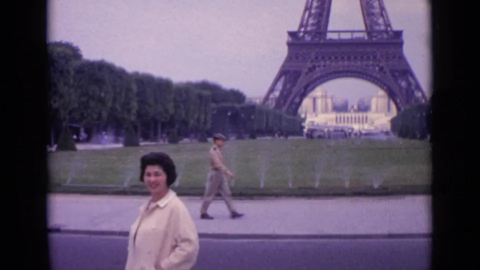 1962 Paris frame with a woman smiling in front of the Eiffel Tower while a uniformed passerby walks across the foreground path.