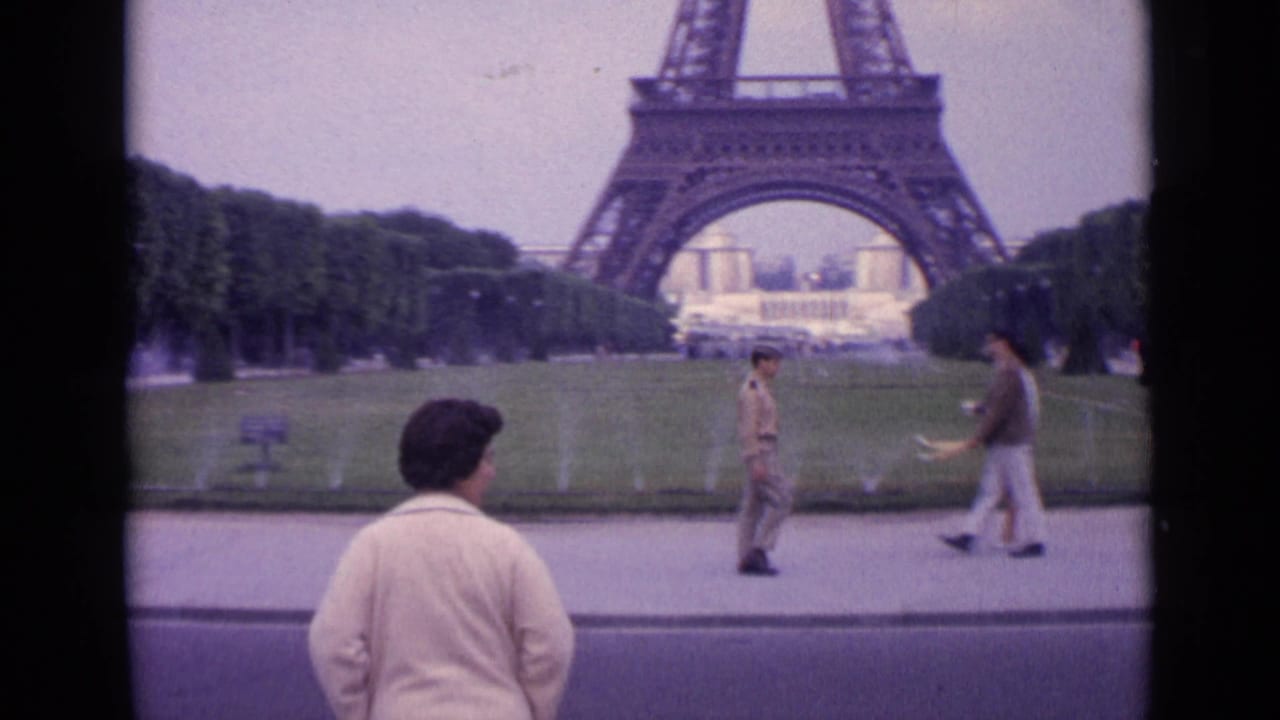 1962 Paris home-movie view of the woman from behind near the Eiffel Tower, with fountains, lawns, and pedestrians crossing the path.
