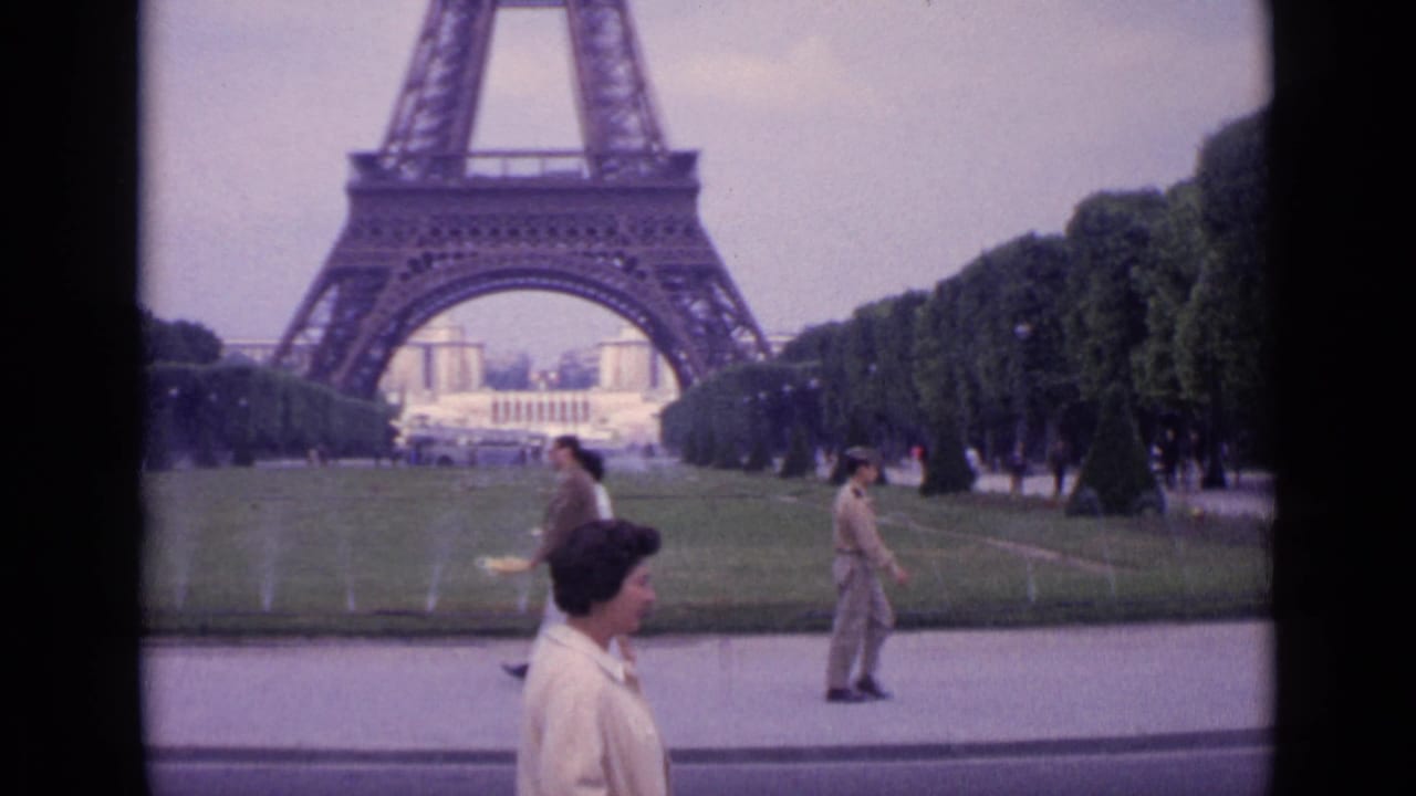 1962 Paris frame showing the woman walking past the camera in front of the Eiffel Tower lawns while other visitors move through the scene.