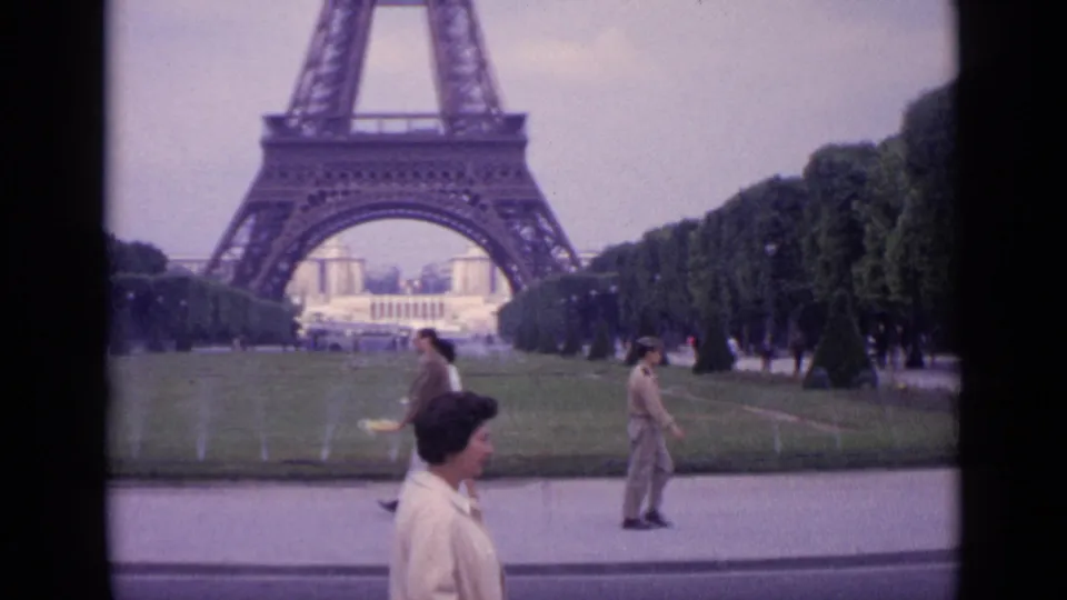1962 Paris frame showing the woman walking past the camera in front of the Eiffel Tower lawns while other visitors move through the scene.