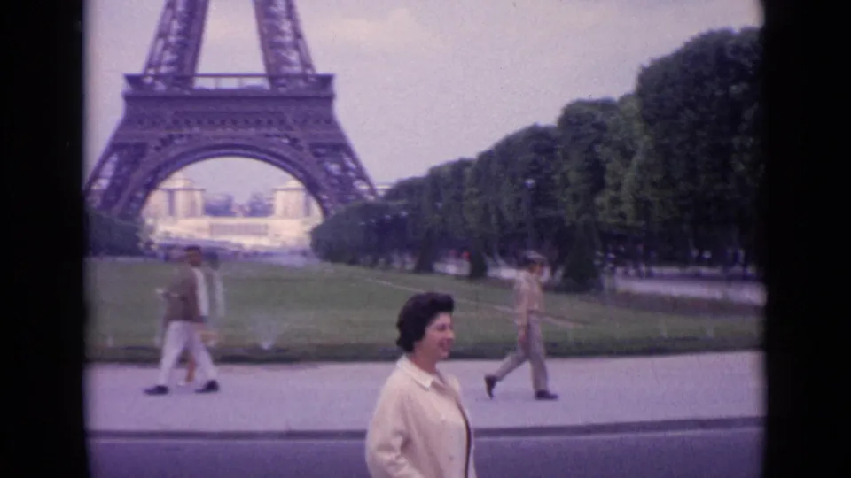 1962 Paris home-movie frame showing a woman walking near lawns with the Eiffel Tower visible in the background.