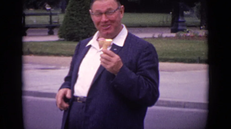 1962 Paris home-movie frame showing a smiling man holding an ice cream cone beside lawns and a public walkway.