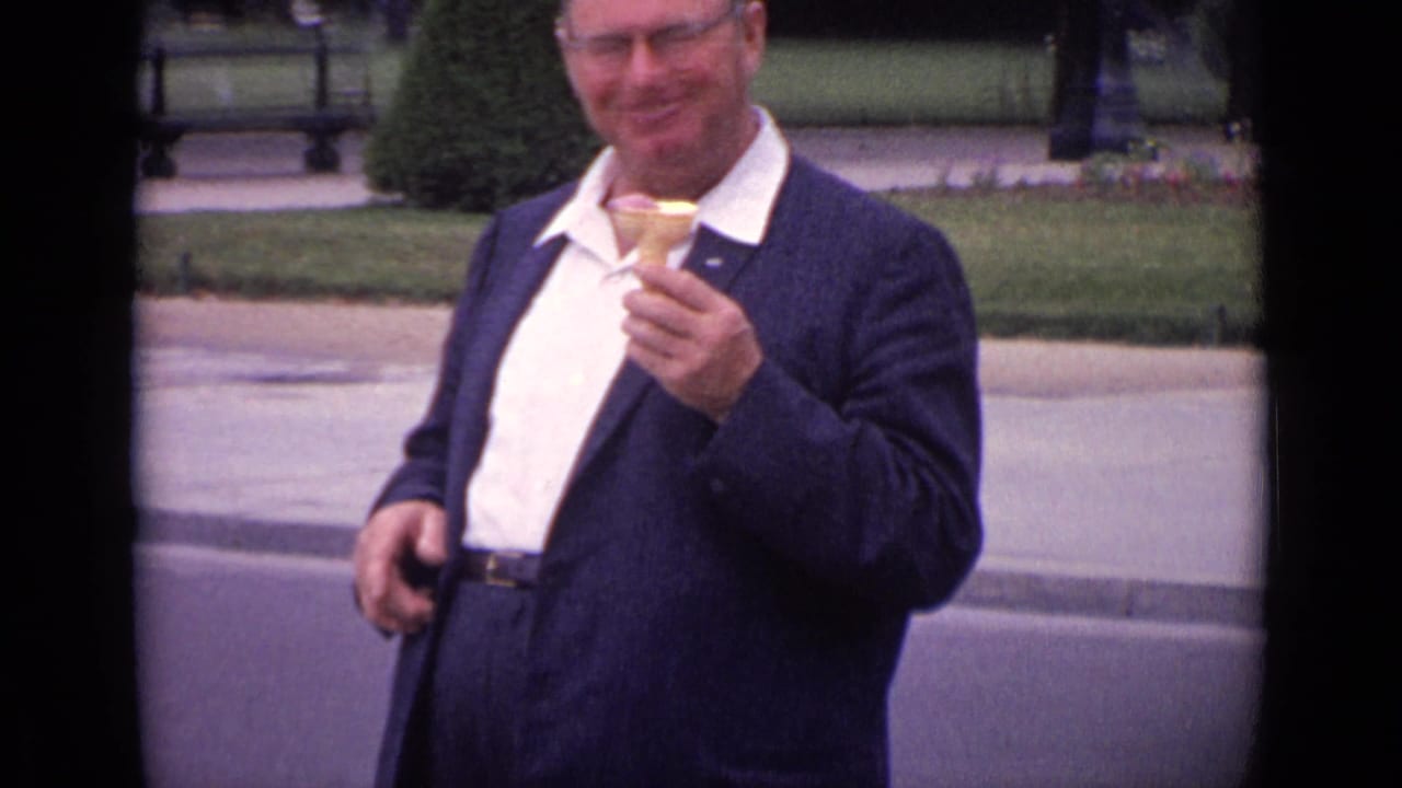 In 1962 Paris home-movie footage, a smiling man in glasses and a dark suit holds an ice cream cone beside formal garden grounds.
