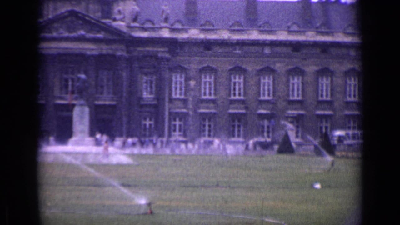 1962 Paris home-movie frame showing a classical building, lawn sprinklers, trees, and a monument with small distant figures.