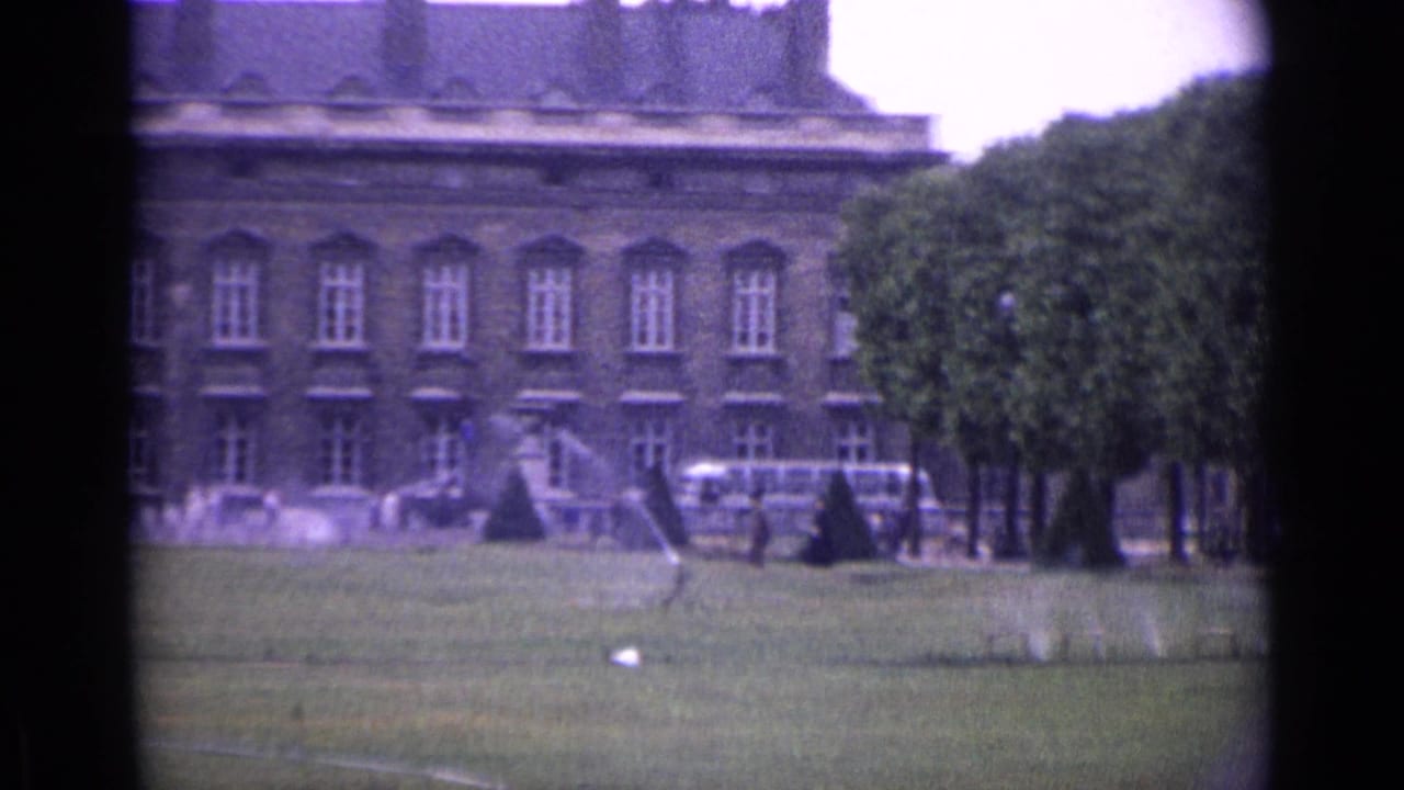 Classical building and lawn with trees, sprinklers, distant people, and vehicles.