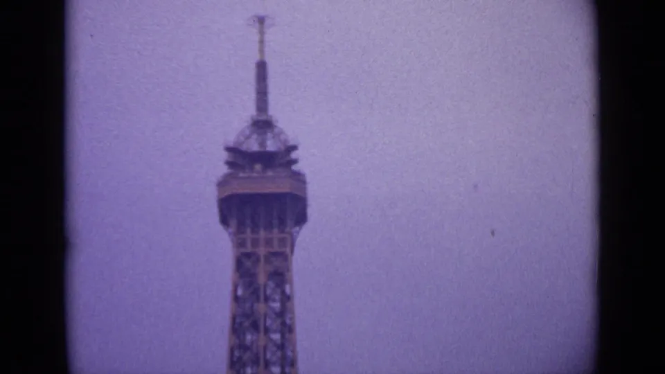 1962 Paris home-movie frame centered on the upper Eiffel Tower, with soft focus and a pale purple-tinged sky.