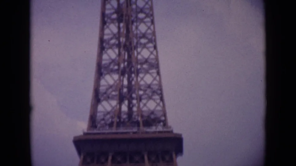 Low-angle 1962 Paris home-movie frame showing the Eiffel Tower base platform, legs, and latticework in faded 8mm color.
