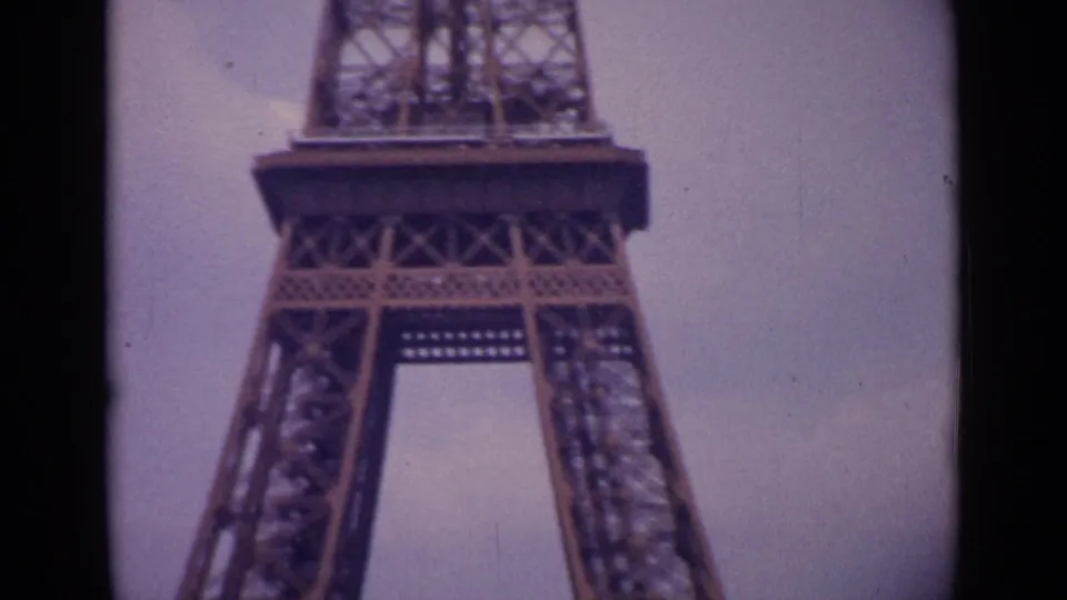 1962 8mm view of the Eiffel Tower base in Paris, showing the lower platform, open arch space, and iron bracing under an overcast sky.