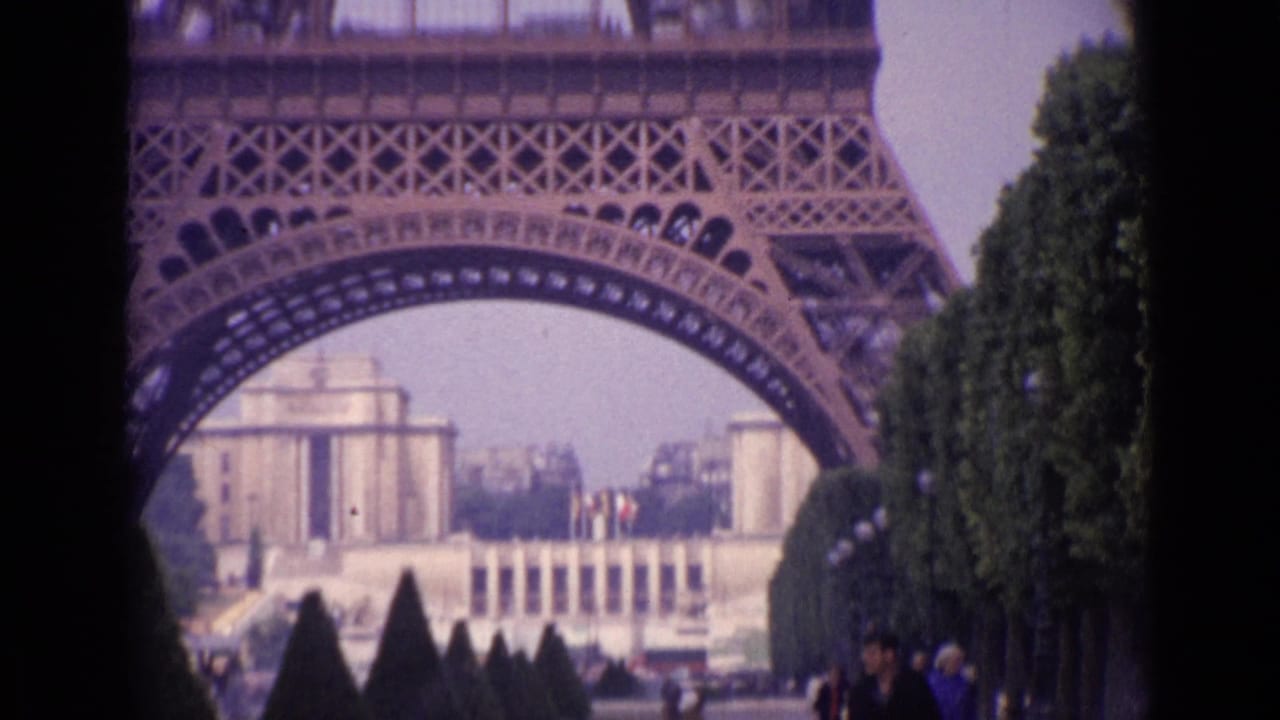 Eiffel Tower arch framing distant buildings and clipped trees with pedestrians at right.