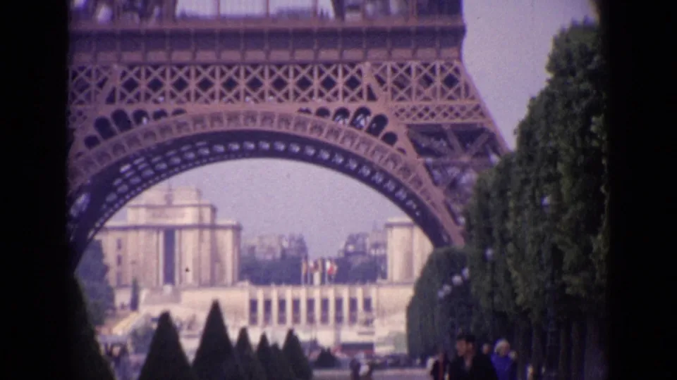 1962 Paris view through the Eiffel Tower arch toward distant Trocadéro buildings, with clipped trees and visitors nearby.