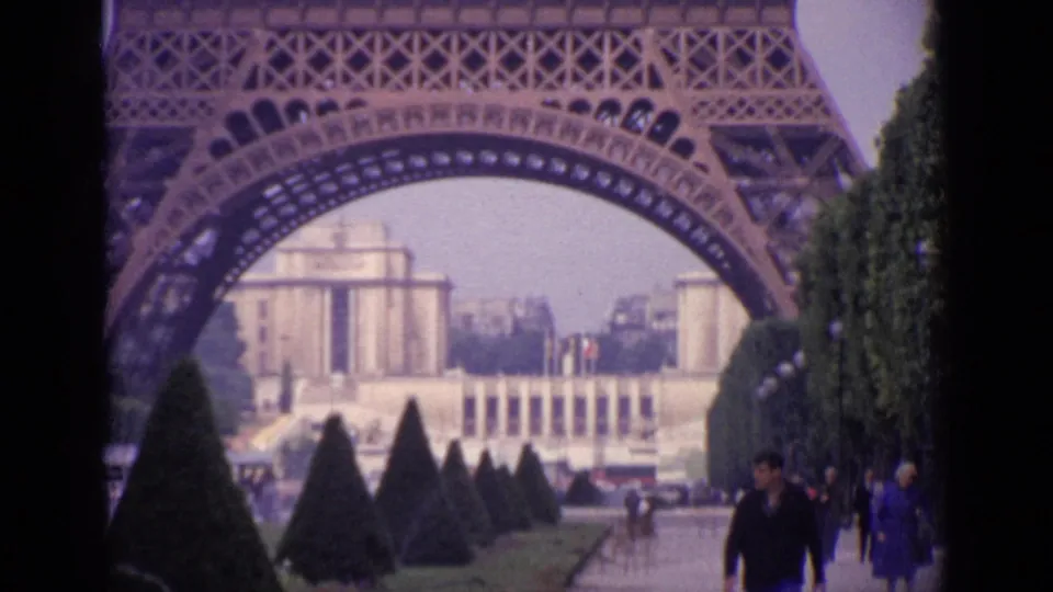 1962 Paris frame with the Eiffel Tower arch, distant Trocadéro architecture, cone-shaped trees, and people walking in the foreground.