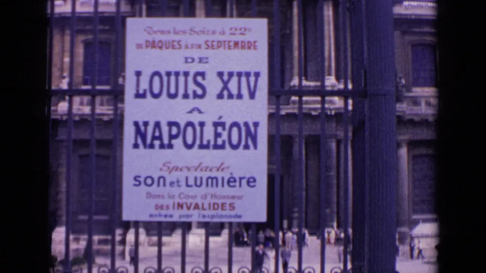 1962 Paris home-movie frame showing a readable French poster for a Louis XIV to Napoléon spectacle on an iron fence near a public building.