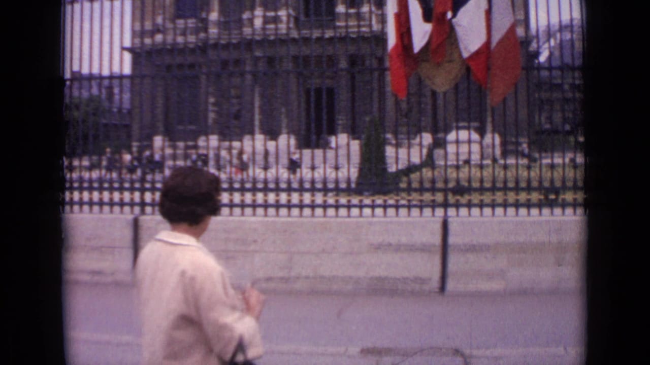 1962 Paris home-movie frame showing a woman near ornate black fencing, with French flags overhead and stone grounds beyond.