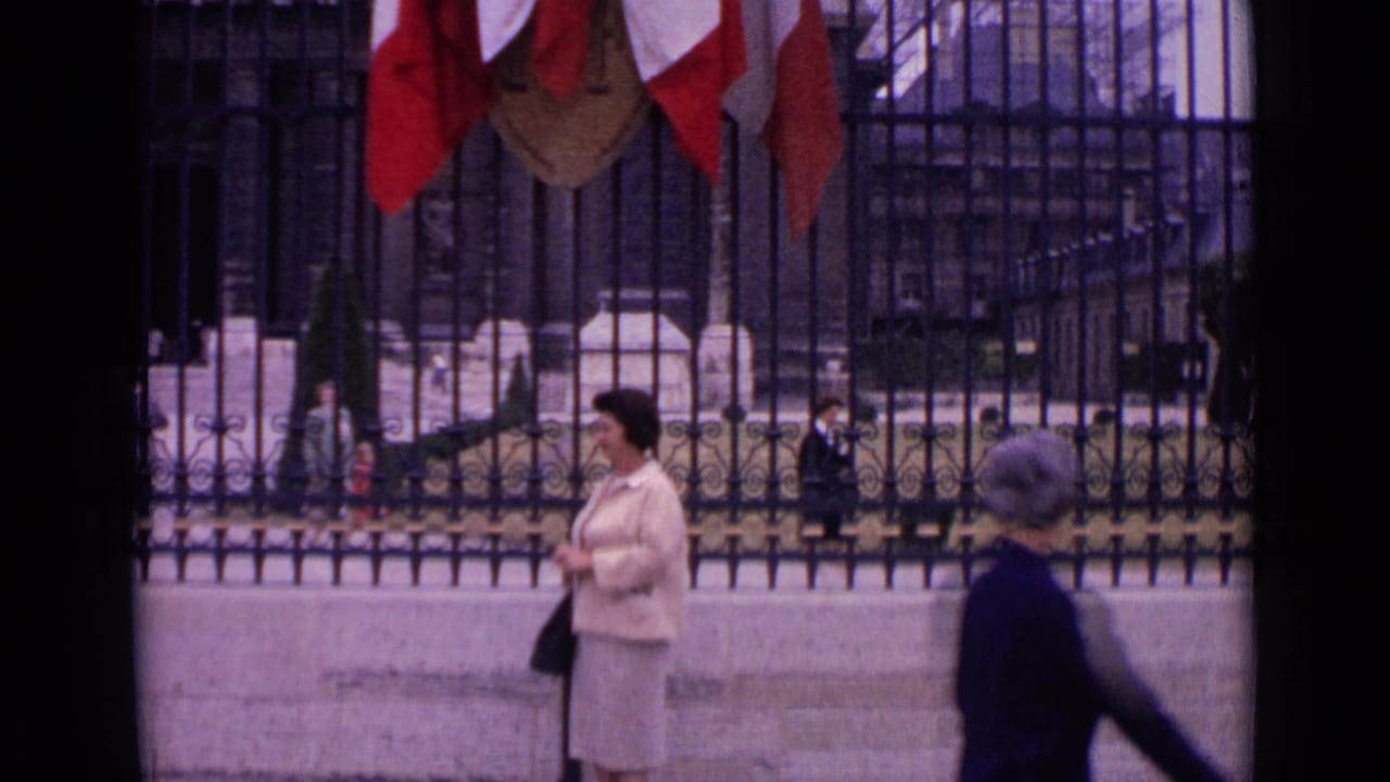 Woman in a light suit stands beside a black fence while another pedestrian passes in front.