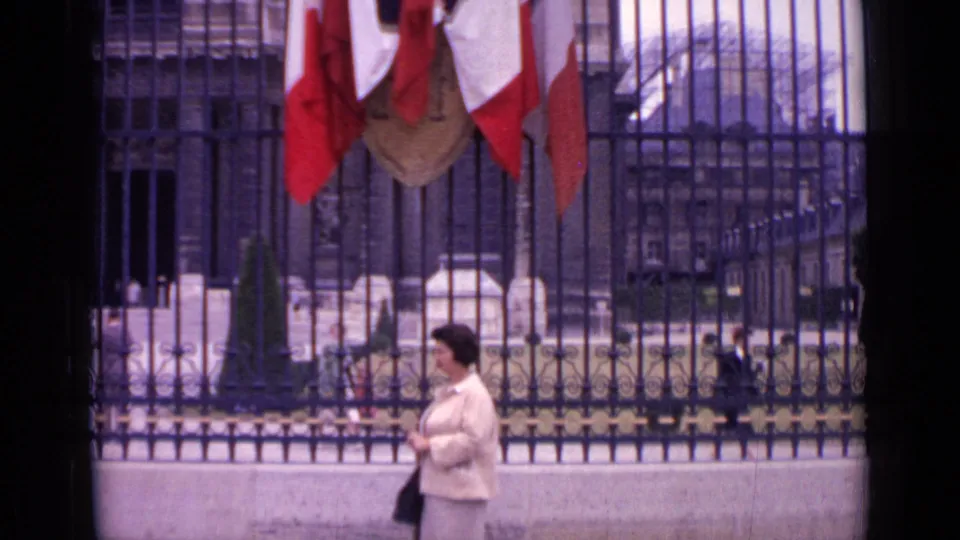 1962 Paris home-movie still of a woman holding a handbag before black railings, with French tricolor flags hanging above the gate.