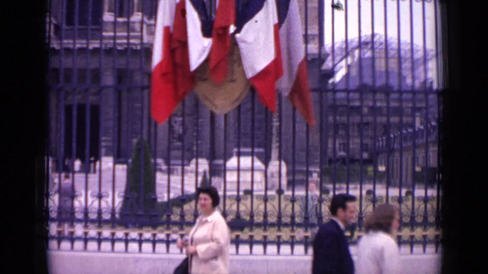 1962 Paris frame showing a woman by the black fence, French flags overhead, and two pedestrians crossing the lower right foreground.