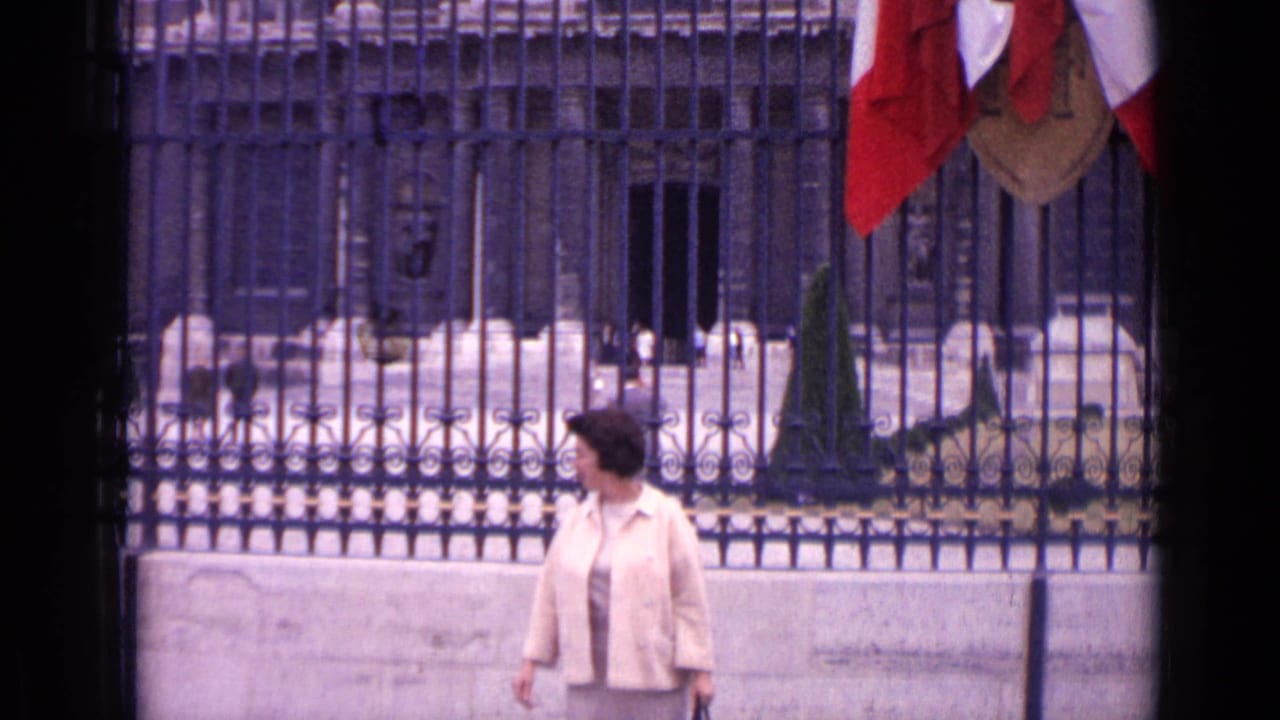 In 1962 Paris home-movie footage, a woman walks beside an ornate iron fence with stone architecture and red and white flags behind her.