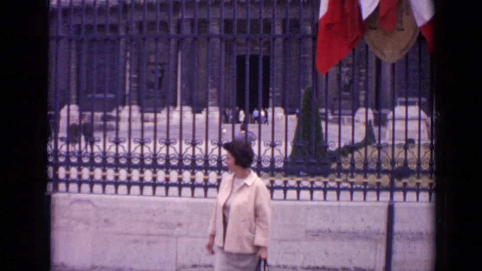 A woman in a light jacket moves along an iron fence in Paris, with formal stone architecture and hanging flags visible in the background.