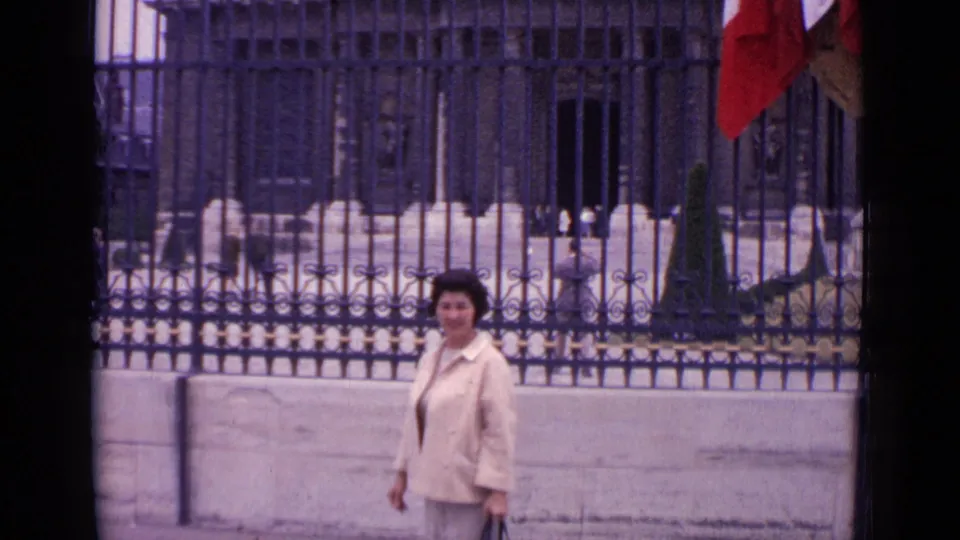 The woman turns toward the camera near ornate Paris railings, with a pale stone wall, flags, and a formal building behind her.