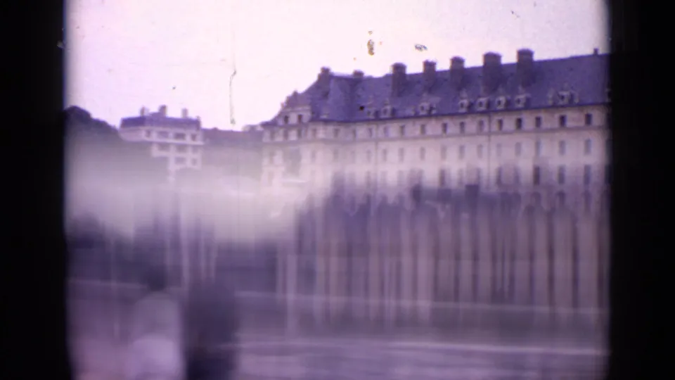 1962 Paris home-movie frame with a long landmark building, dark roof, stone wall, and heavy motion blur from a moving vehicle.